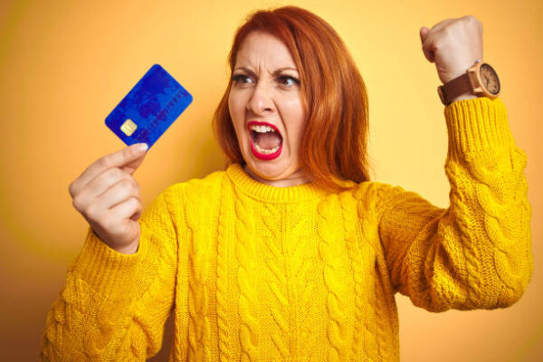 Young customer redhead woman holding credit card over yellow isolated background annoyed and frustrated shouting with anger, crazy and yelling with raised hand, anger concept